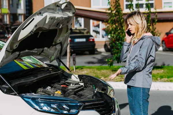 Upset young woman talking by smartphone and looking at broken car — Stock Photo