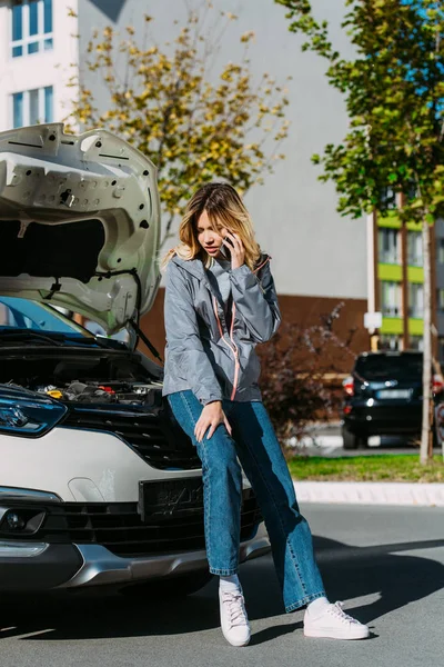 Mujer joven molesta hablando en el teléfono inteligente mientras está sentado en el coche roto - foto de stock