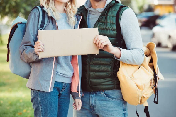 Cropped shot of young couple with backpacks holding blank card on road — Stock Photo