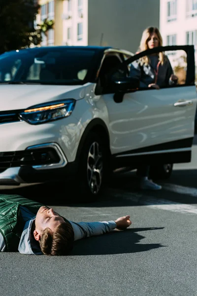 Injured young man lying on road and woman standing near car behind after traffic collision — Stock Photo