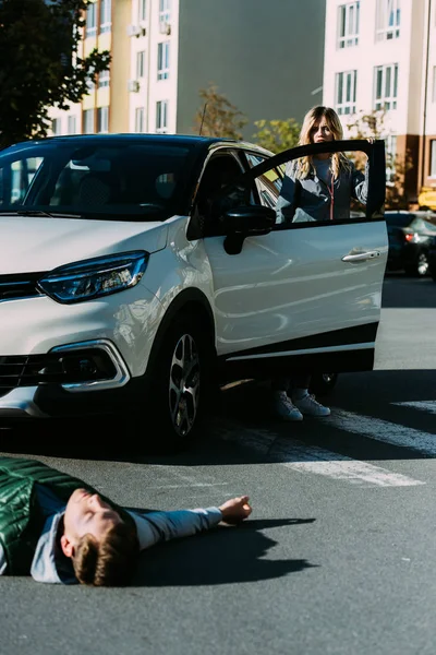 Mujer abriendo la puerta del coche y mirando al hombre herido acostado en la carretera después de accidente de tráfico - foto de stock