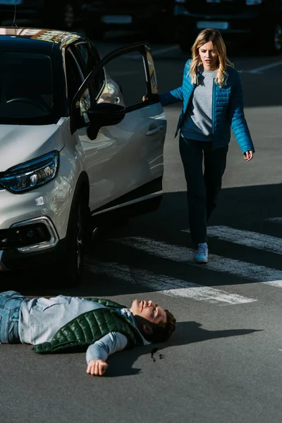 Scared woman opening car door and going to injured young man lying on road after traffic accident — Stock Photo
