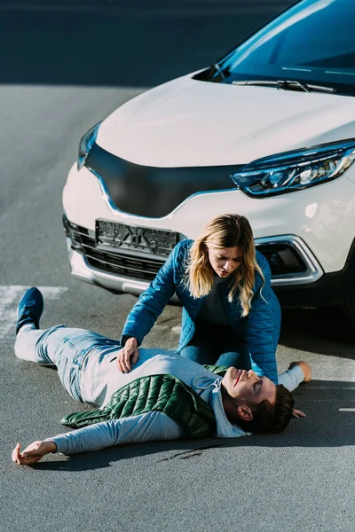 Vista de ángulo alto de la mujer joven mirando tocando hombre herido acostado en la carretera después de accidente de tráfico - foto de stock