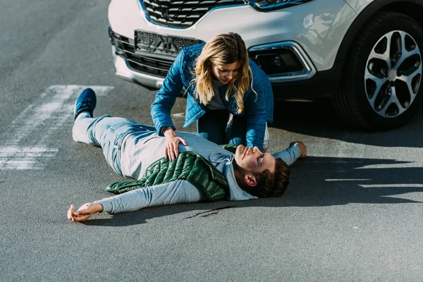 Vista de ángulo alto de la mujer joven asustada tocando hombre herido acostado en la carretera después de accidente de coche - foto de stock