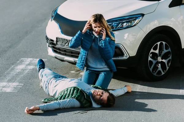 Vista de ángulo alto de la mujer joven asustada mirando al hombre herido y llamando de emergencia después de un accidente de coche - foto de stock