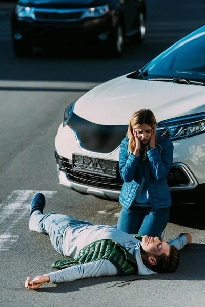 Високий кут зору на налякану молоду жінку, яка дивиться на жертву, що лежить на дорозі після автомобільної аварії — стокове фото
