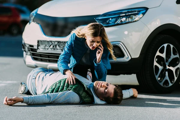 Scared young woman calling emergency and looking at injured man on road after traffic accident — Stock Photo