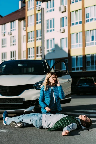 Junge Frau ruft Notruf und kniet nach Verkehrsunfall neben verletztem Mann — Stockfoto