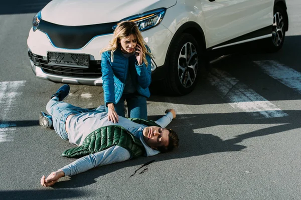 Vista ad alto angolo della giovane donna spaventata che chiama l'emergenza e tocca l'uomo ferito sulla strada dopo l'incidente stradale — Foto stock