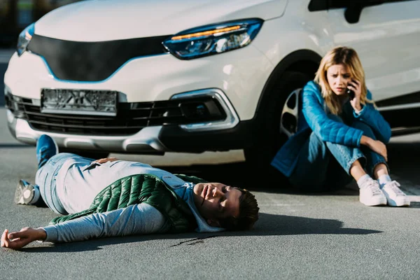 Mujer joven llamando de emergencia mientras el hombre herido que yace en la carretera después de la colisión de vehículos de motor - foto de stock