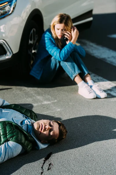 Visão de alto ângulo da jovem mulher assustada chamando emergência enquanto homem ferido deitado na estrada após colisão de trânsito — Fotografia de Stock