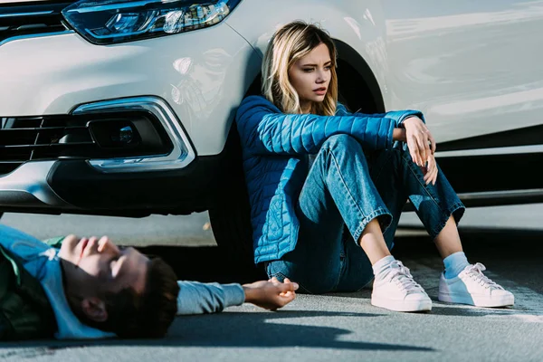 Mujer joven asustada sentada cerca de coche y hombre herido acostado en la carretera después de la colisión de tráfico - foto de stock