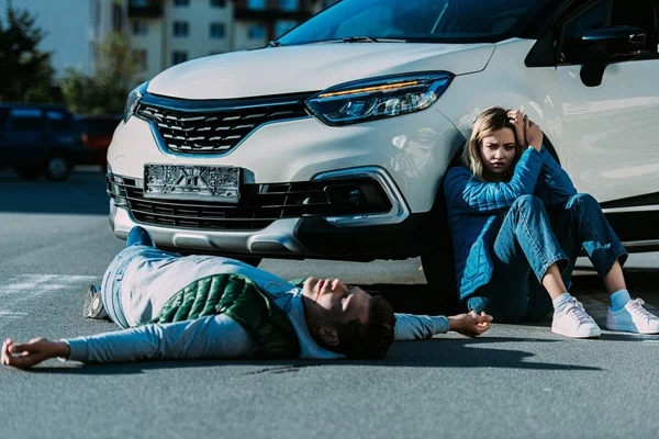 Scared young woman sitting near car and looking at injured man lying on road after traffic collision — Stock Photo