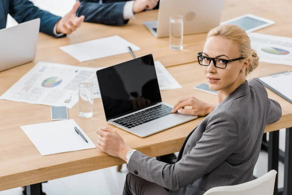 Jeune femme dans des lunettes assis à table avec ordinateur portable dans le bureau d'assurance et regardant la caméra — Photo de stock