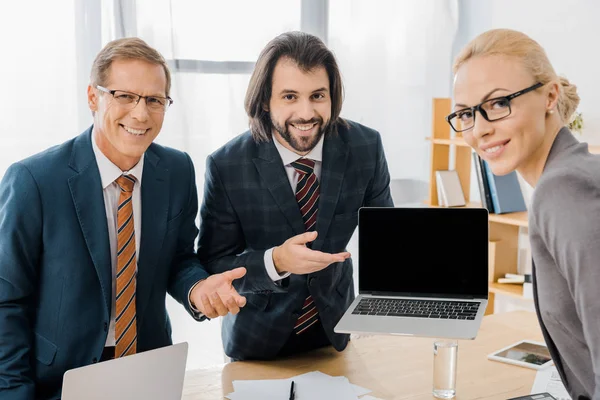 Young smiling male insurance agent standing with workers and pointing at laptop in office — Stock Photo