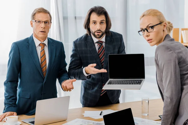 Young serious male insurance agent standing with workers and pointing at laptop in office — Stock Photo