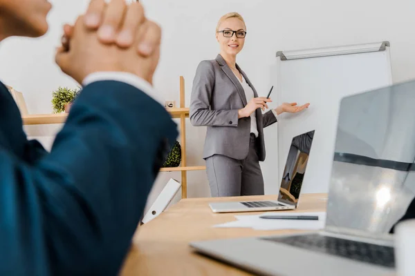 Jeune femme souriante pointant vers le tableau blanc et l'homme assis à la table dans le bureau — Photo de stock