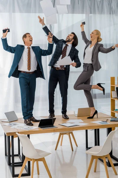 Alegres trabajadores de seguros bailando sobre la mesa y tirando papeles en la reunión en la oficina - foto de stock