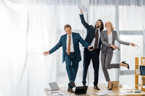 Fröhliche Versicherungsangestellte, die auf dem Tisch tanzen und bei einem Treffen im Büro mit Papieren werfen — Stockfoto