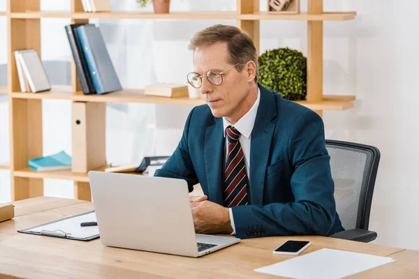 Adult serious businessman in glasses sitting at table and using laptop in office — Stock Photo