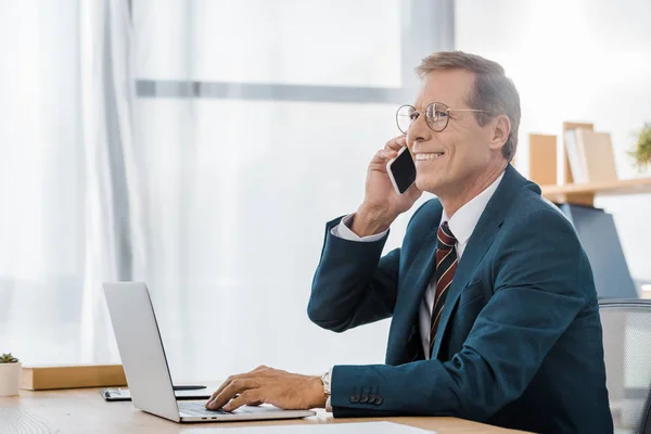 Взрослый улыбающийся бизнесмен в очках разговаривает на смартфоне и использует ноутбук в офисе — стоковое фото