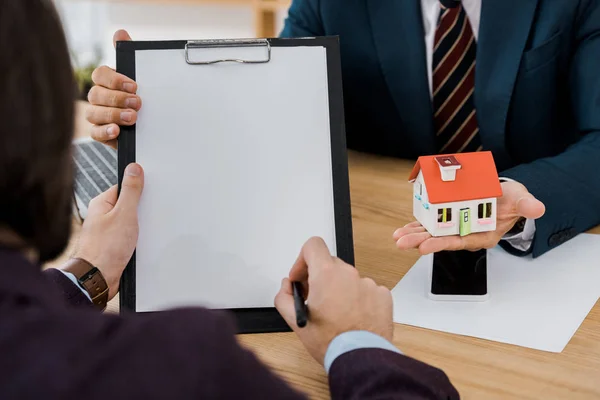 Man signing contract on clipboard while insurance agent holding house model — Stock Photo