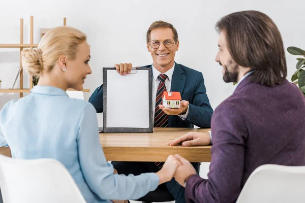 Glückliches Paar hält Händchen, während Versicherungsvertreter Vertrag und kleines Haus zeigt — Stockfoto