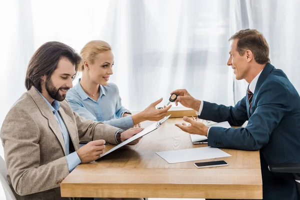 Agente de seguros dando la llave a la mujer mientras que el hombre firma papeles - foto de stock