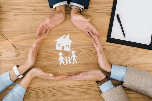 Gente haciendo círculo con las manos en la mesa de madera con casa de papel y la familia en el interior, seguro de vida - foto de stock