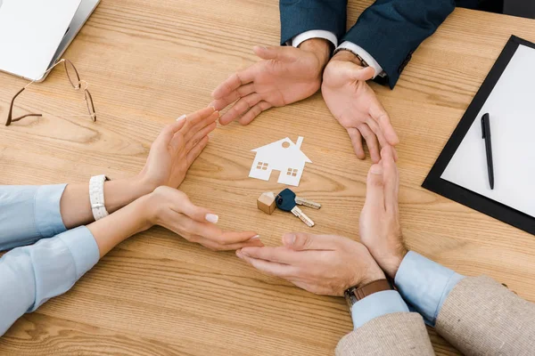 Gente haciendo círculo con las manos en la mesa de madera con casa de papel y llaves en el interior, seguro de casa - foto de stock