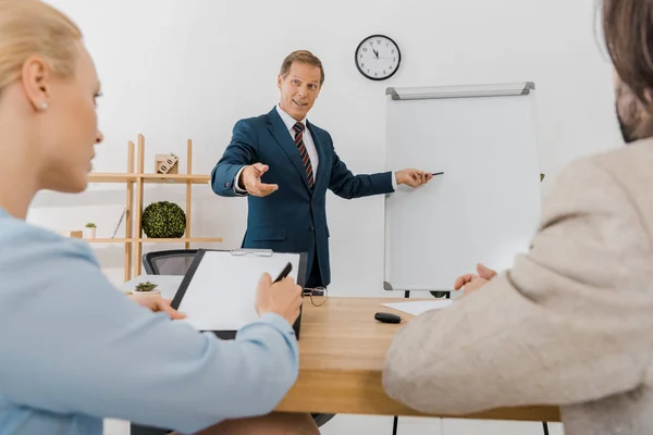 Geschäftsmann zeigt auf Whiteboard, während Paar Vertrag auf Klemmbrett unterzeichnet — Stockfoto