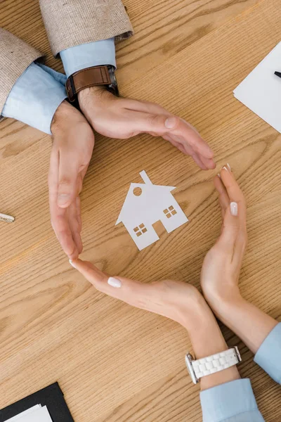 Pareja haciendo círculo con las manos en la mesa de madera con casa de papel en el interior, seguro de casa - foto de stock