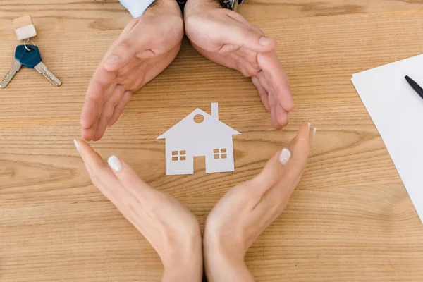 Pareja haciendo círculo con las manos con la casa de papel blanco en el interior de la mesa de madera, seguro de casa - foto de stock