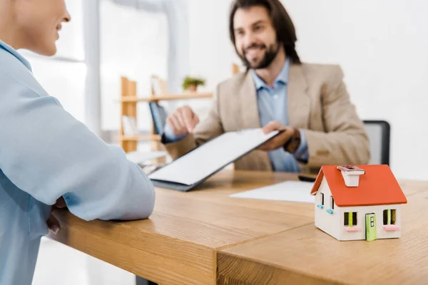 Lächelnder Mann zeigt Frau Versicherungsvertrag mit Hausmodell auf Holztisch — Stockfoto