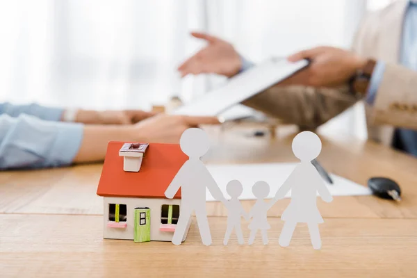 Libro bianco taglio famiglia e modello di casa su tavolo in legno con persone offuscate sullo sfondo, vita e assicurazione casa — Foto stock