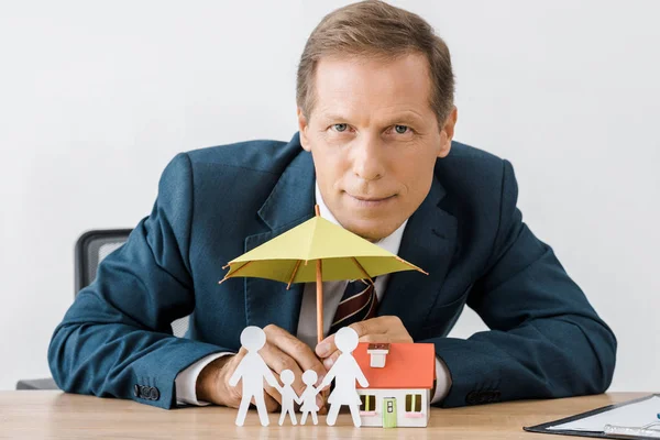 Homme d'affaires avec famille de coupe de papier, modèle de maison et parapluie sur table en bois — Photo de stock