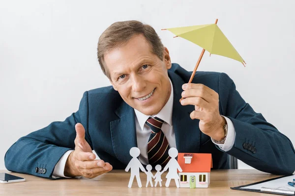 Hombre de negocios que sostiene el paraguas con la familia de corte de papel y el modelo de casa en la mesa de madera - foto de stock