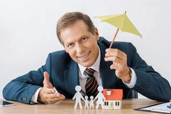 Businessman holding umbrella with paper cut family and house model on wooden table — Stock Photo