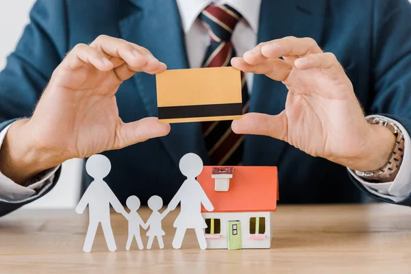 Männliche Hände halten Kreditkarte mit Hausmodell und papiergeschnittener Familie auf Holztisch — Stockfoto