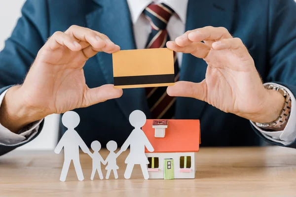 Manos masculinas que sostienen la tarjeta de crédito con el modelo de casa y la familia de papel cortado en mesa de madera - foto de stock