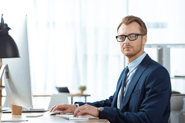 Selbstbewusster Geschäftsmann mit Brille blickt in die Kamera und arbeitet am Tisch mit Computermonitor im modernen Büro — Stockfoto