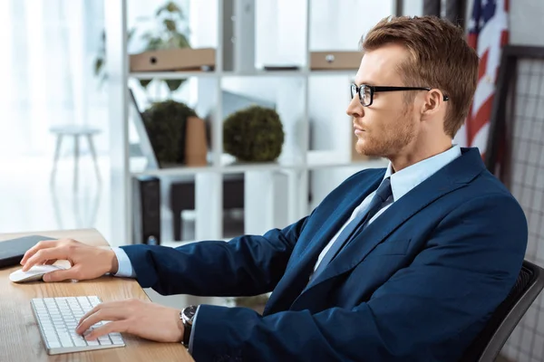Vista lateral de hombre de negocios guapo en gafas que trabajan en la mesa con el teclado del ordenador y el ratón en la oficina moderna - foto de stock