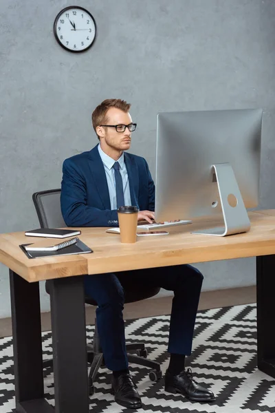 Seriöser junger Geschäftsmann mit Brille am Tisch mit Computermonitor im modernen Büro — Stockfoto