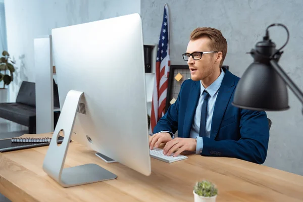 Alegre hombre de negocios en las gafas que trabajan a la mesa con el monitor de ordenador en la oficina moderna - foto de stock