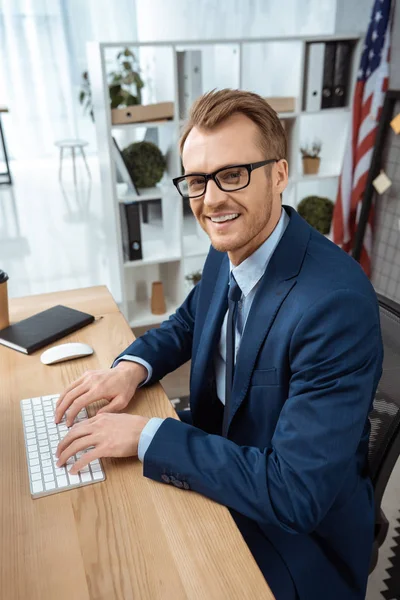 Усміхнений бізнесмен в окулярах дивиться на камеру і друкує на комп'ютерній клавіатурі за столом в сучасному офісі — стокове фото