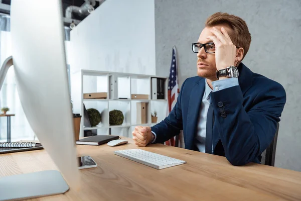 Selective focus of tired businessman in eyeglasses gesturing by hand at table with computer monitor in office — Stock Photo