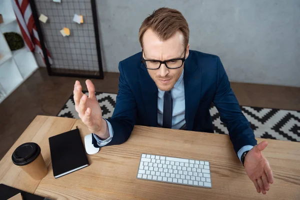 Высокий угол зрения раздраженного бизнесмена в очках жестикулируя руками за столом с компьютерной клавиатурой и мышью в офисе — стоковое фото