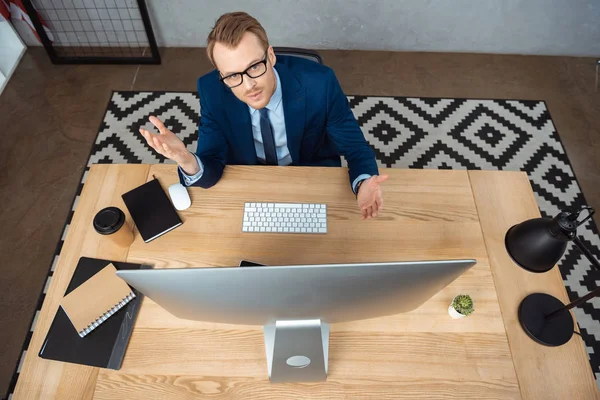 Overhead veiw of businessman in eyeglasses  looking at camera and gesturing by hands at table with computer monitor in office — Stock Photo