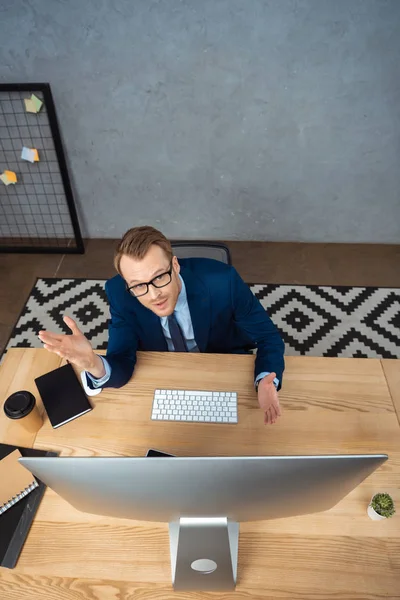 Высокий угол обзора молодого бизнесмена в очках жестом руками за столом с компьютерным монитором в офисе — стоковое фото