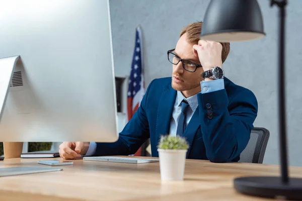 Втомлений бізнесмен в окулярах працює за столом з комп'ютерним монітором в сучасному офісі — стокове фото